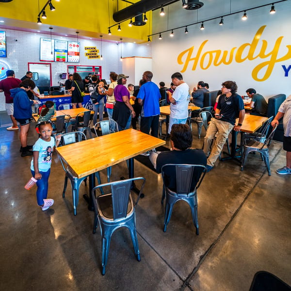 Customers gather in a brightly lit Howdy Hot Chicken restaurant with a large yellow "Howdy" sign.