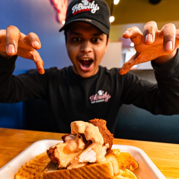 A man in a Howdy Hot Chicken hat and shirt excitedly reaches for a fried chicken sandwich.