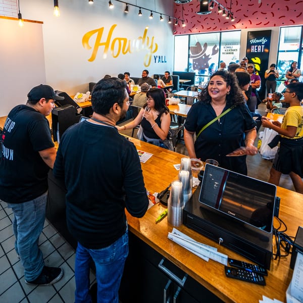 Customers and staff interact at the counter of Howdy Hot Chicken, with a busy dining area in the background.