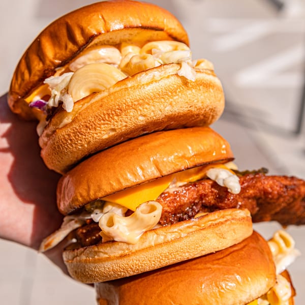 A hand holds a tall stack of three Howdy Hot Chicken sandwiches with mac and cheese.